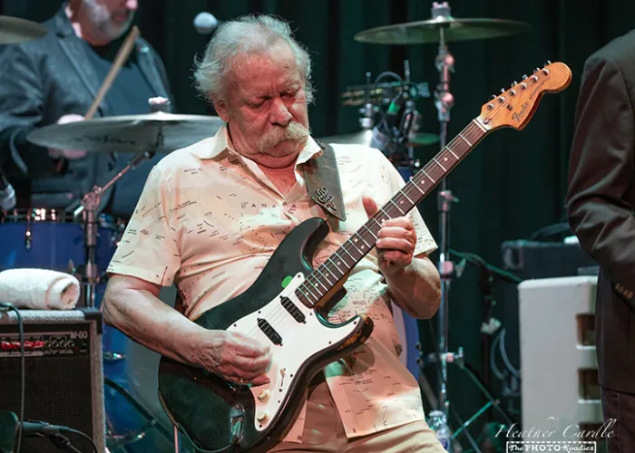 A man playing an electric guitar on stage with a set of drums in the background