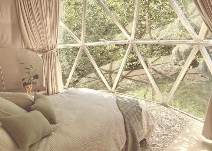 A soft sunlit room inside a glamping dome with triangular glass windows and bed displayed with earthy tones of green and beige. 