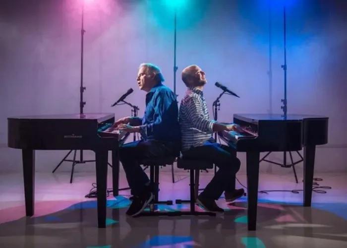 Two people facing away from each other singing and playing piano
