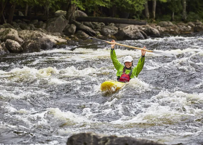 Woman in yellow kayak, mid-river holding paddle in the air