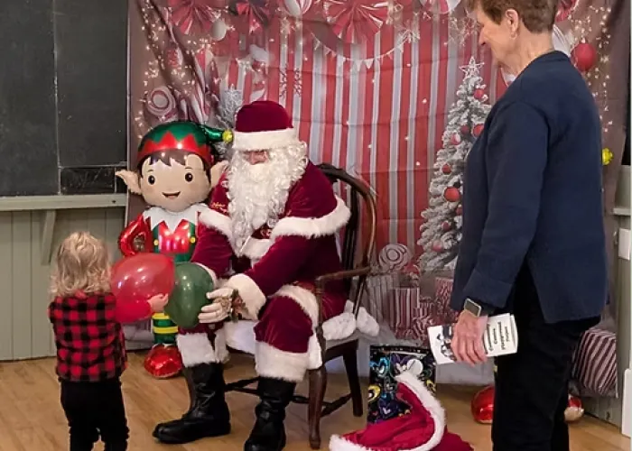 Photo of child holding a balloon out to santa while santa sits and holds a balloon in the child's direction, christmas background of red and white colours, adult stands watching