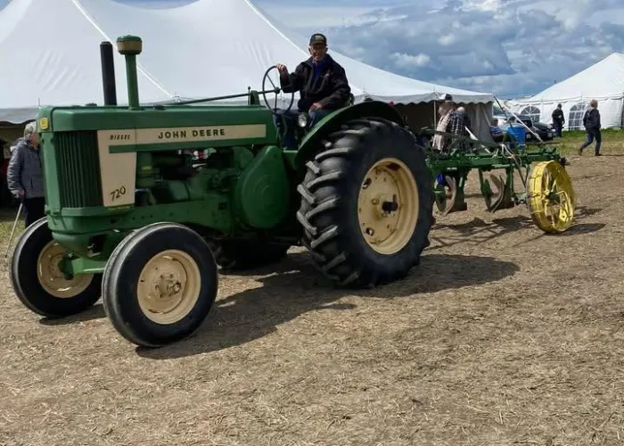 Person driving green and yellow John Deere tractor, large white tent in background, blue skies, white clouds