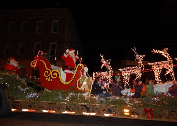 Photo at night of santa in sleigh on parade float with lit up reindeer and children sitting in front