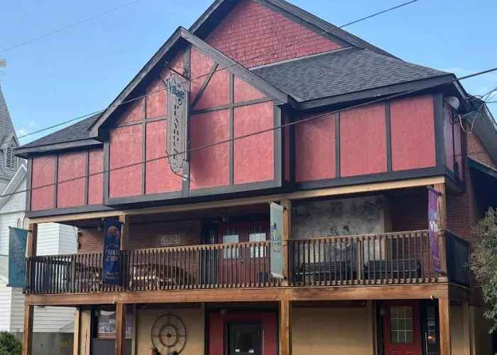 Photo of the Bancroft Village Playhouse, red top storey, flag with name of venue, balcony on second storey, porch with beige background on first storey, blue skies behind