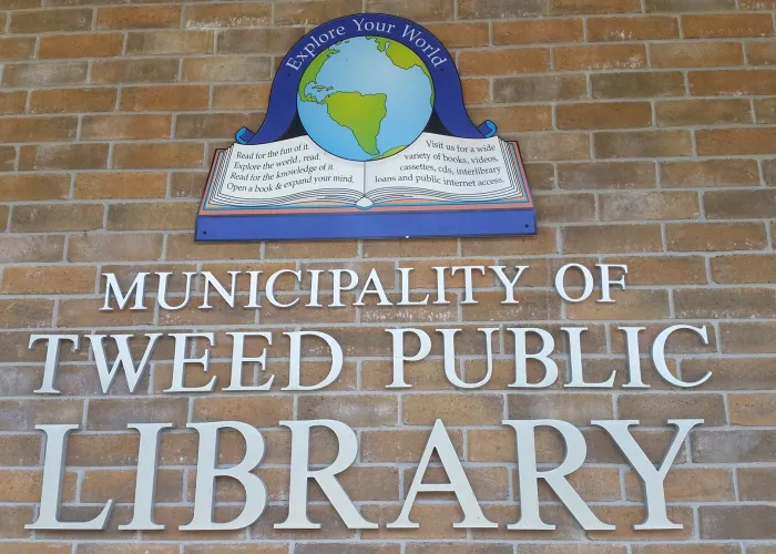 Logo of Tweed Public Library of open book with earth sitting in centre of pages, name of library beneath all on a brick wall